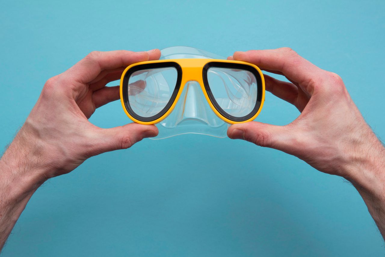 A close-up of hands holding a yellow snorkeling mask against a bright blue background.